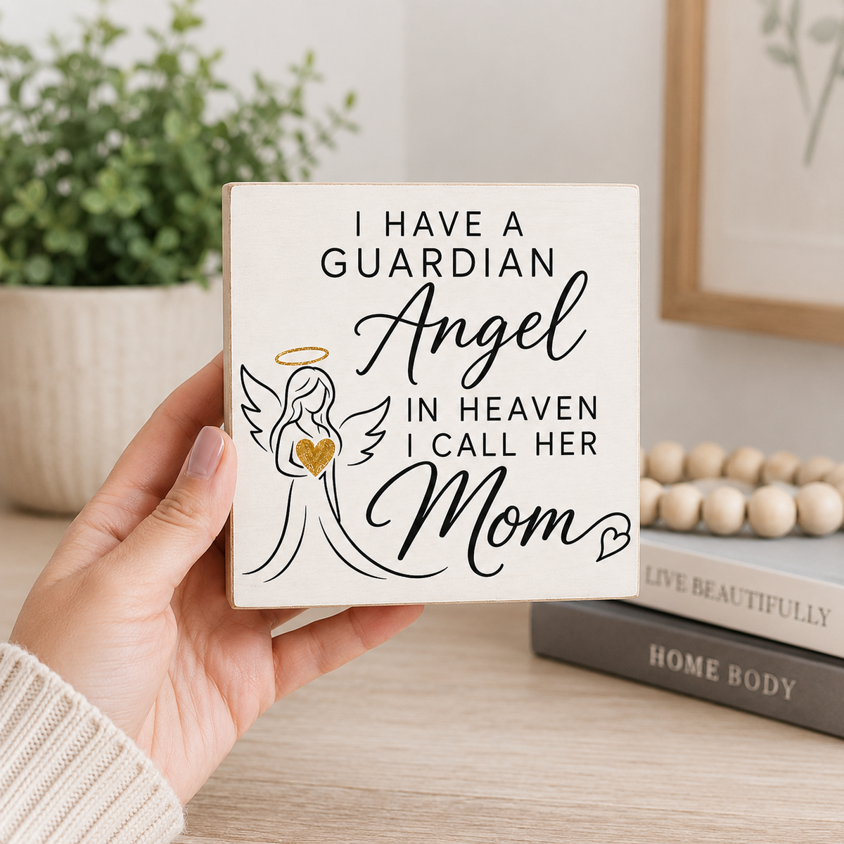 A hand holds a square cream-colored wooden sign with the printed message 'I HAVE A GUARDIAN Angel IN HEAVEN I CALL HER Mom' alongside a simple angel illustration. The background shows a softly blurred home interior with a potted plant, books, and a wooden bead garland.
