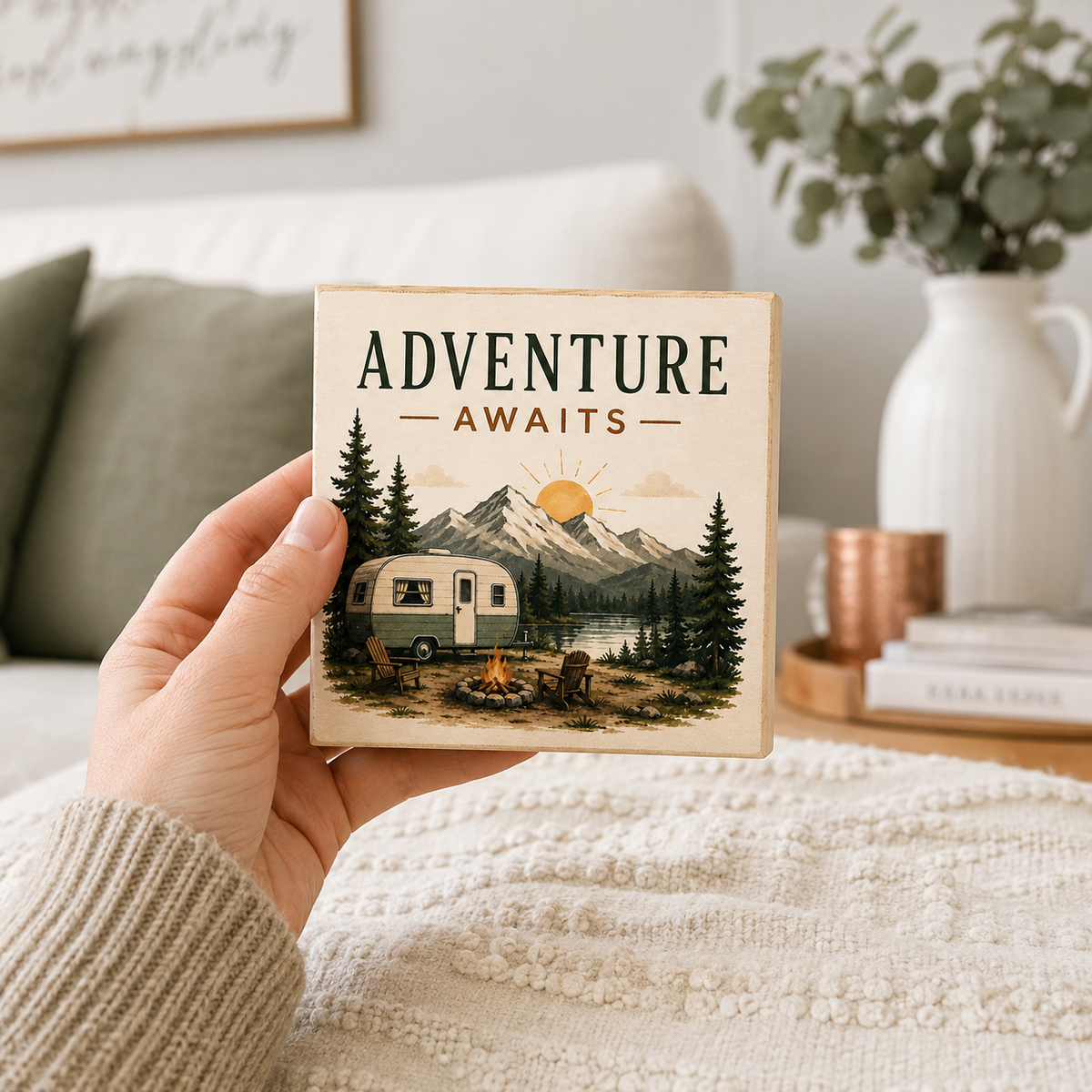 A hand holds a square coaster featuring a retro camping scene with the text 'ADVENTURE AWAITS' above an illustration of a vintage camper, mountains, trees, and a campfire. The background shows a cozy living room setting with a sofa, blanket, and decorative vase.