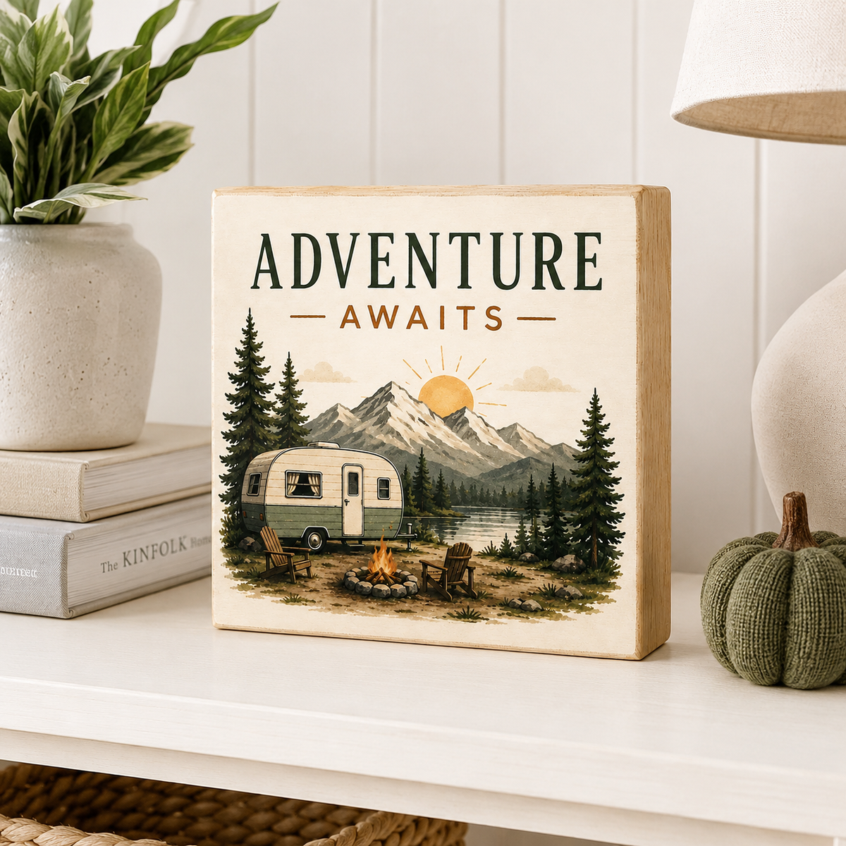 A square wooden sign with a vintage-style camping illustration and the text 'ADVENTURE AWAITS' stands on a white shelf. The sign features a retro camper, mountains, pine trees, and a campfire.