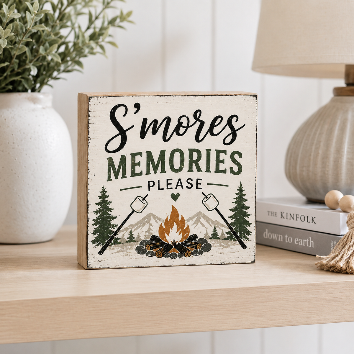 A square wooden sign with a distressed cream background featuring the text 'S'mores MEMORIES PLEASE' above an illustration of a campfire, trees, mountains, and marshmallows. The sign is styled on a light wood shelf next to a white vase with greenery, a lamp, and books.