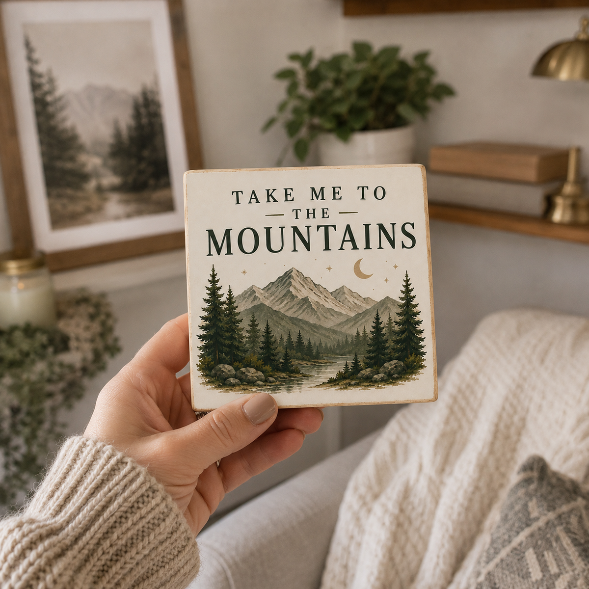A hand wearing a beige knit sweater holds a square ceramic tile coaster featuring a scenic mountain and pine forest illustration with the text 'TAKE ME TO THE MOUNTAINS'. The background shows a cozy living room setting with a sofa, a framed print, and a plant.