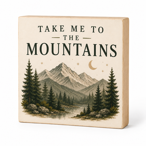 A square wooden block sign with a light natural wood frame and a cream-colored face, printed with the text 'TAKE ME TO THE MOUNTAINS' above an illustrated mountain landscape with pine trees, stars, and a crescent moon.