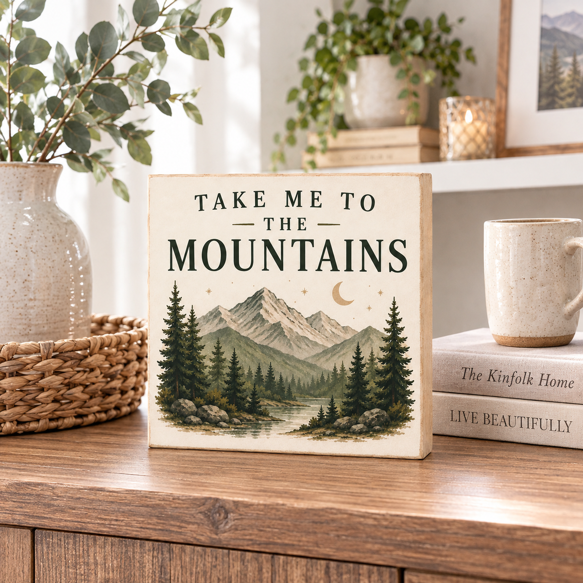 A square decorative sign with the phrase “TAKE ME TO THE MOUNTAINS” and an illustrated mountain landscape, displayed on a wooden table in a styled home setting with books, a mug, and plants.