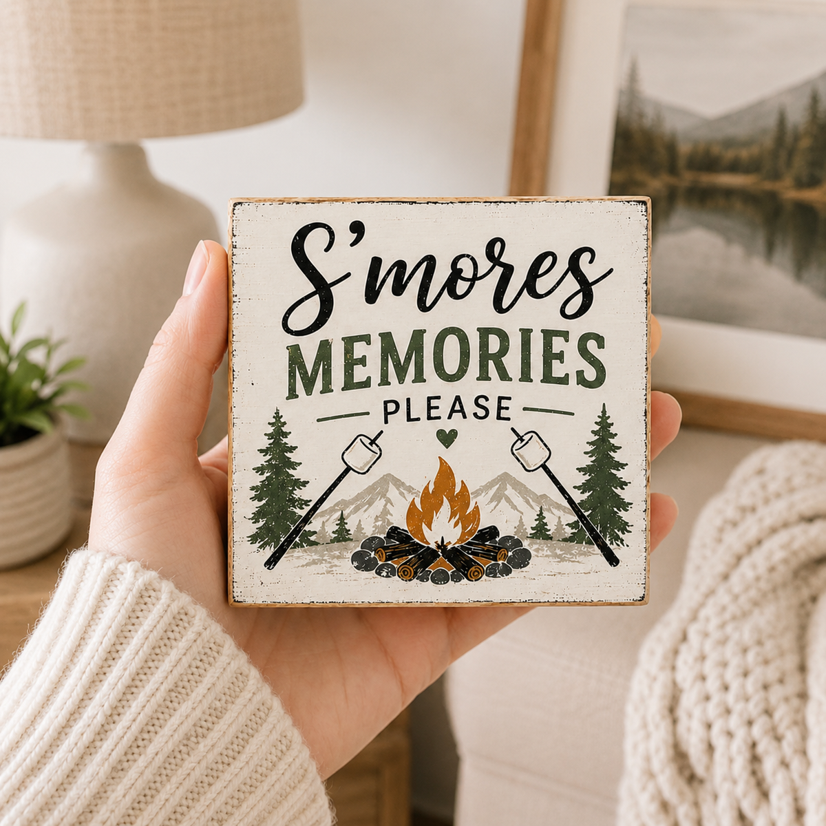 A hand holds a square, rustic white wooden sign that reads 'S'mores Memories Please' with an illustration of a campfire, marshmallows, pine trees, and mountains. The background shows a cozy indoor setting with a lamp, framed picture, and knit sweater.