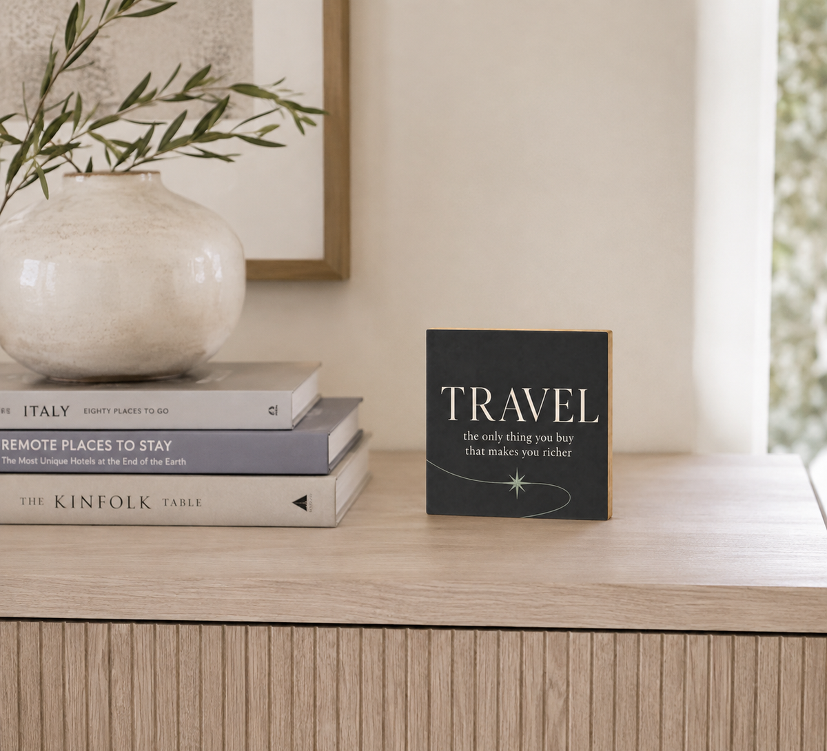 A small square black decorative sign with the word 'TRAVEL' in large cream letters and a small star graphic, standing on a light wood console table beside stacked books and a white vase with greenery in a softly lit interior.
