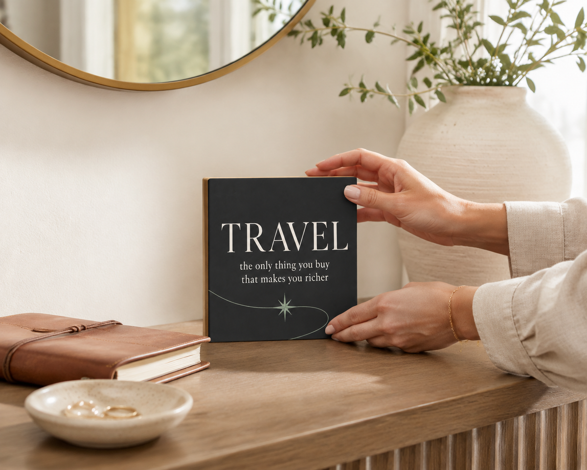 A person's hands are shown holding a black hardcover journal on a wooden console table. The journal features the word 'TRAVEL' in white serif lettering with the subtitle 'the only thing you buy that makes you richer' in a smaller font.
