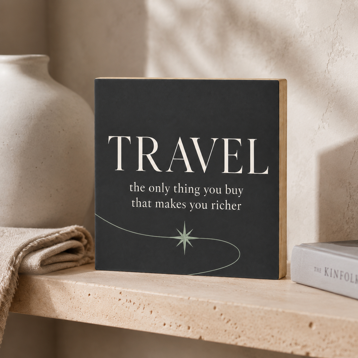 A square decorative book or plaque with a dark charcoal cover stands on a light stone shelf. The cover reads “TRAVEL” in large white serif letters, with the subtitle “the only thing you buy that makes you richer” below, plus a pale mint line drawing.