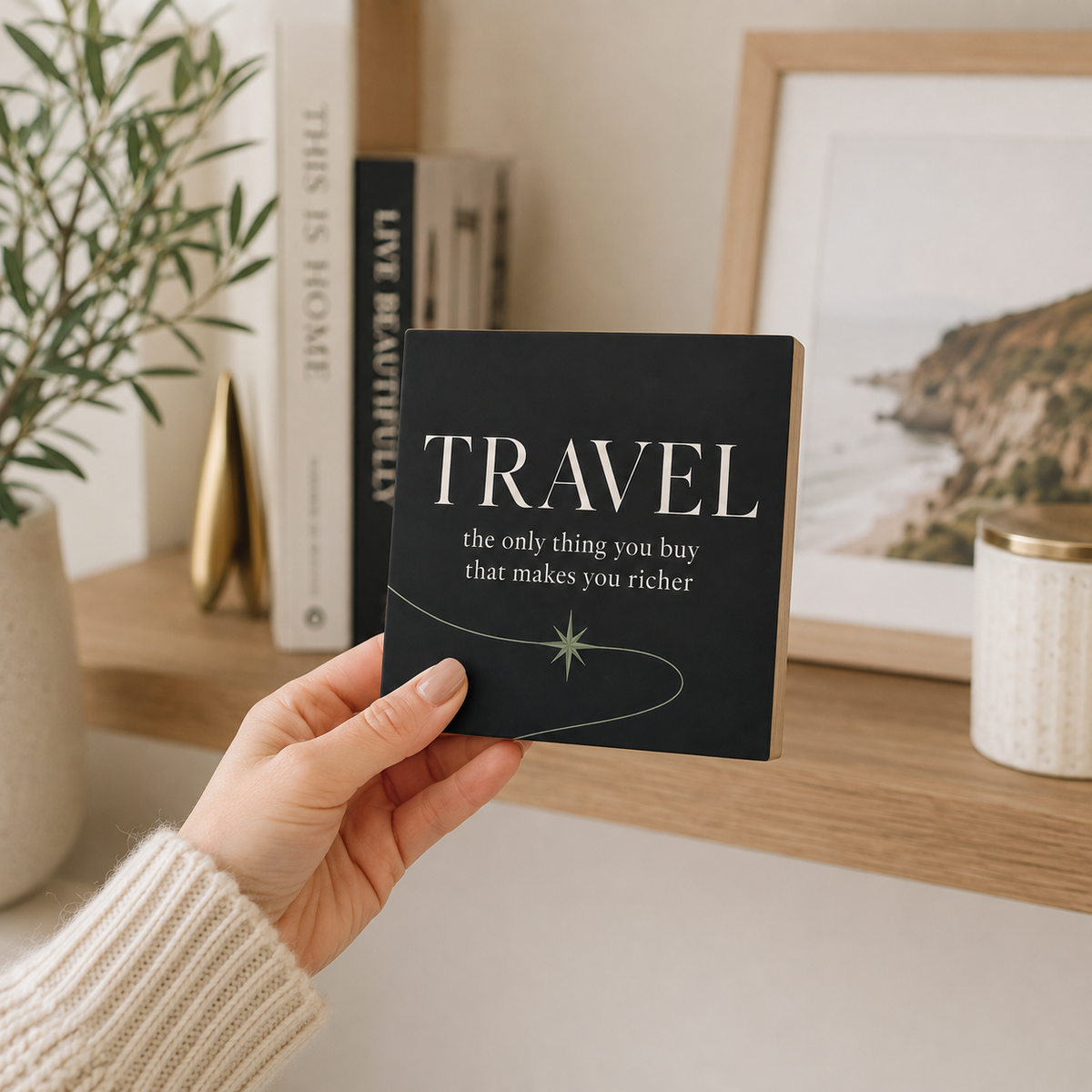A hand in a cream knit sweater holds a square black card or coaster featuring the word 'TRAVEL' in large white letters with the phrase 'the only thing you buy that makes you richer' below it. The background shows a warm, styled interior shelf with books, a plant, a candle, and framed wall art.