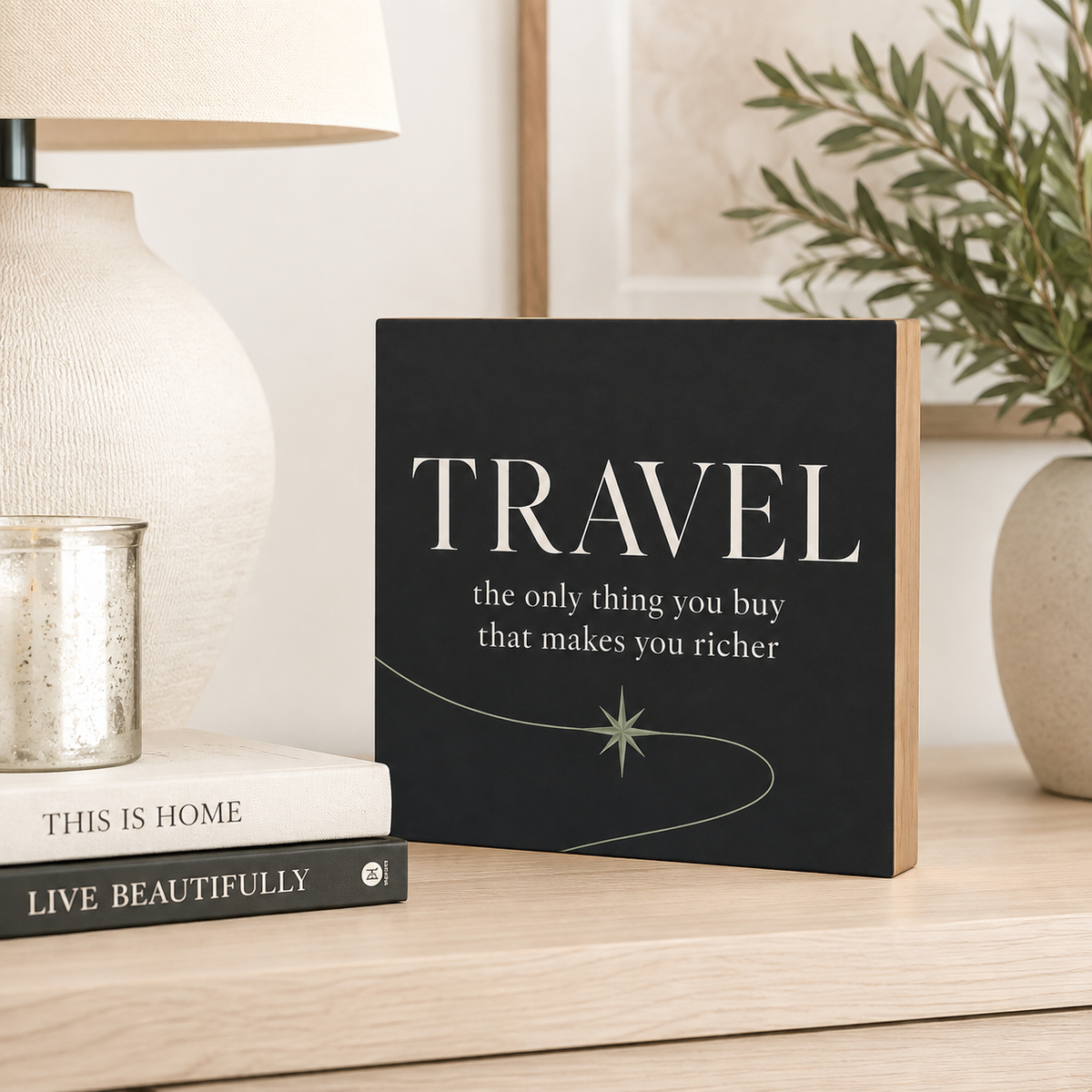 A square decorative sign with a black background and white text reading “TRAVEL the only thing you buy that makes you richer,” styled on a light wood surface beside a lamp, books, and a plant.