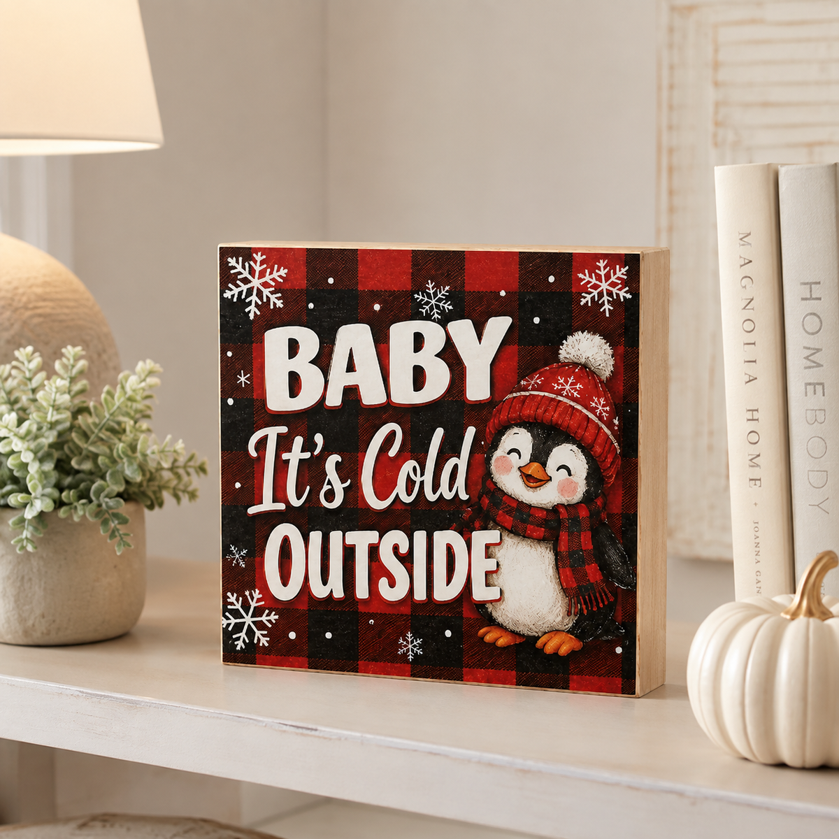A square decorative sign with a red and black buffalo plaid pattern, featuring the text 'BABY It's Cold OUTSIDE' in white lettering and an illustration of a penguin wearing a red hat and scarf. The sign is styled on a white shelf next to a potted plant, books, and a small pumpkin.