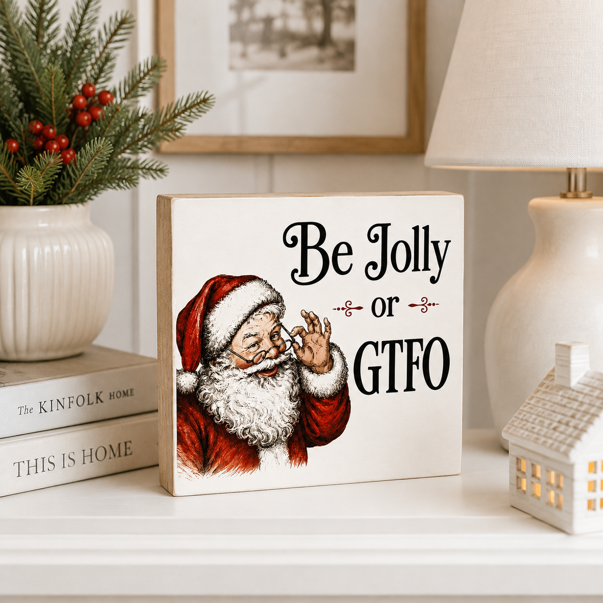 A square decorative sign featuring an illustrated Santa Claus and the text 'Be Jolly or GTFO' in black script, styled on a white shelf beside a lamp, a small house decor, books, and a potted evergreen.