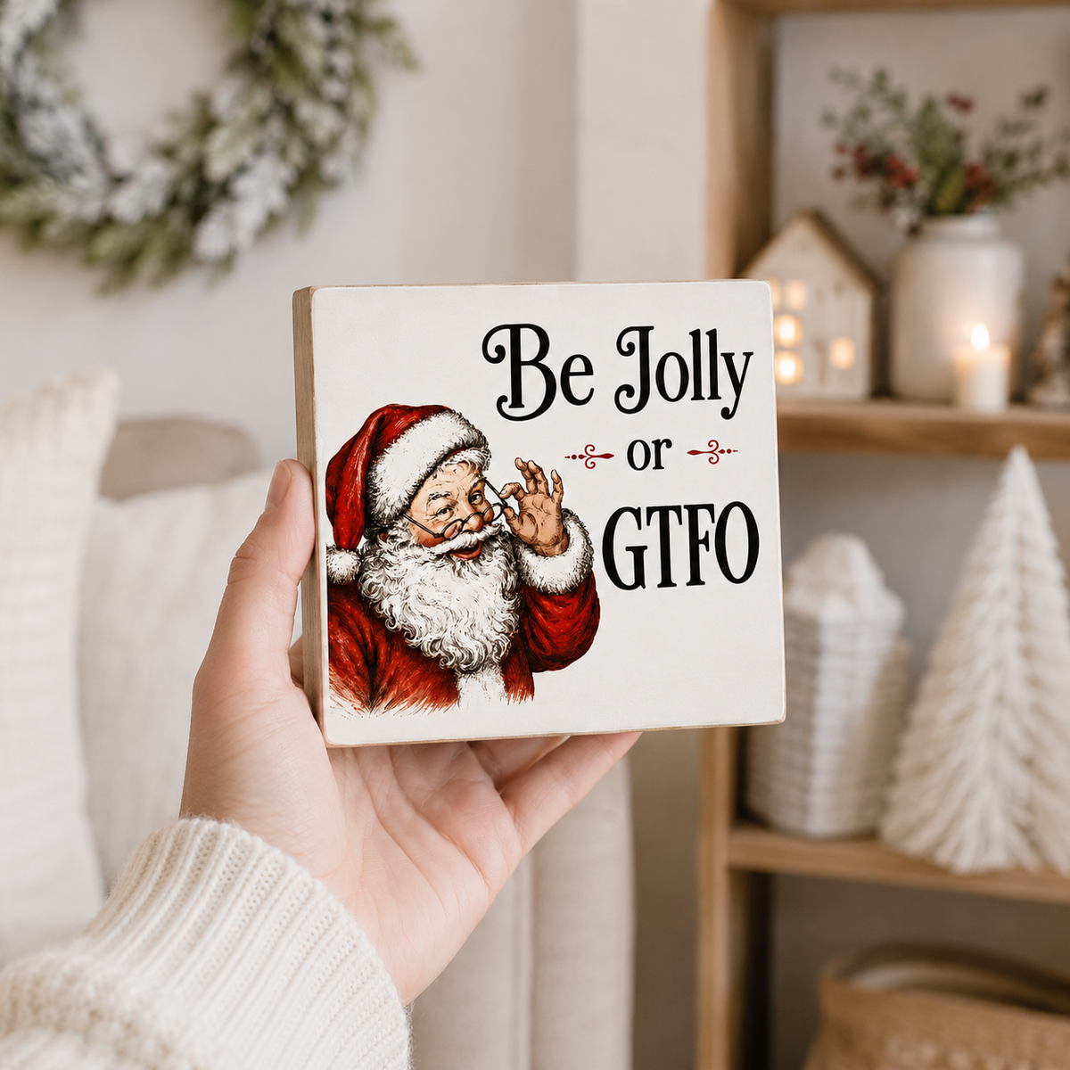 A hand holds a square wooden sign with a vintage-style illustration of Santa and the text 'Be Jolly or GTFO' in a cozy, decorated home setting.