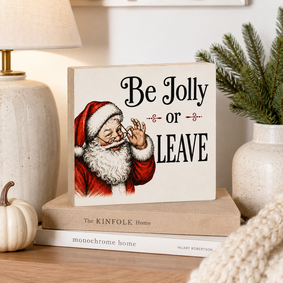 A square decorative sign with a vintage-style illustration of Santa and the text 'Be Jolly or LEAVE' sits on stacked books on a wooden surface. The scene is styled with a lamp, a small pumpkin, greenery, and a knit blanket in a warm indoor setting.
