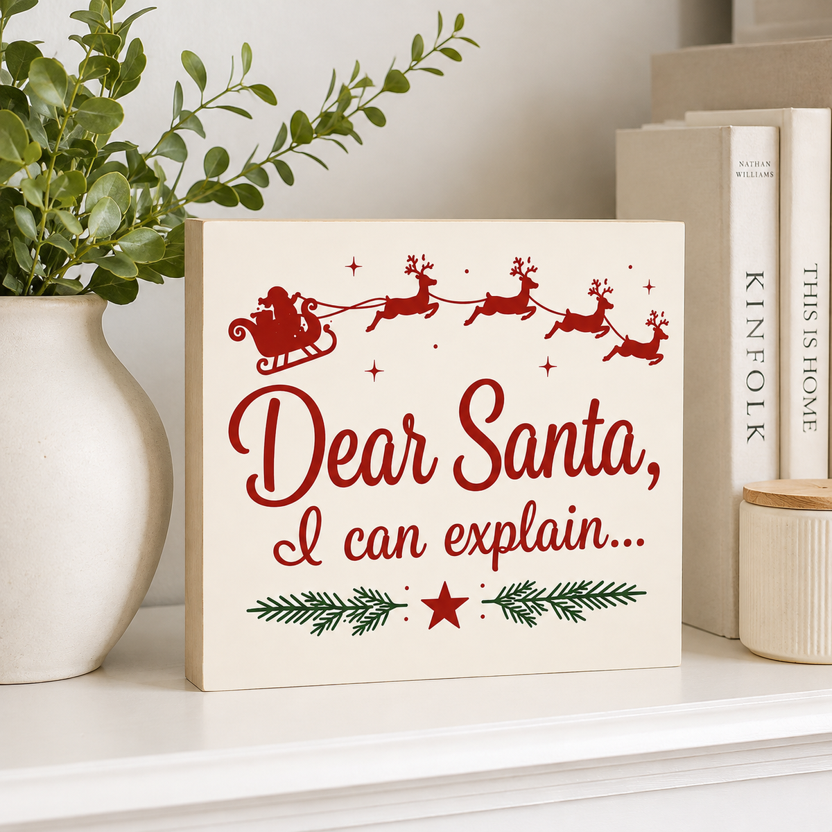 A square cream-colored Christmas sign with red and green printed holiday text and illustrations, displayed on a white shelf next to a white vase with green leaves and books.