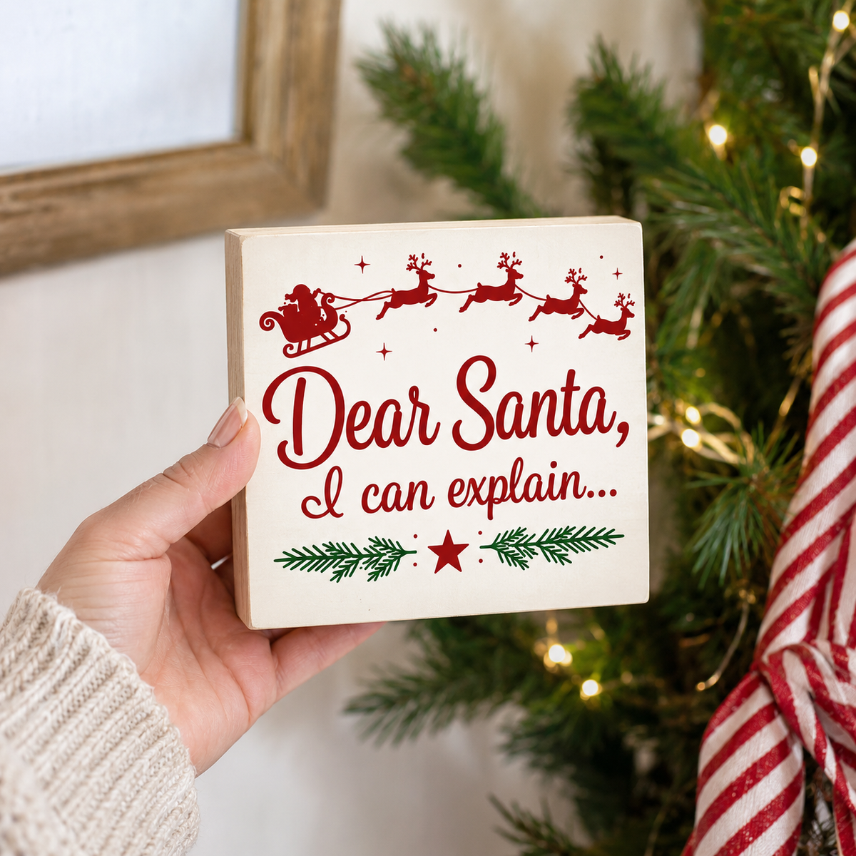 A hand holds a square wooden sign with a red 'Dear Santa, I can explain.