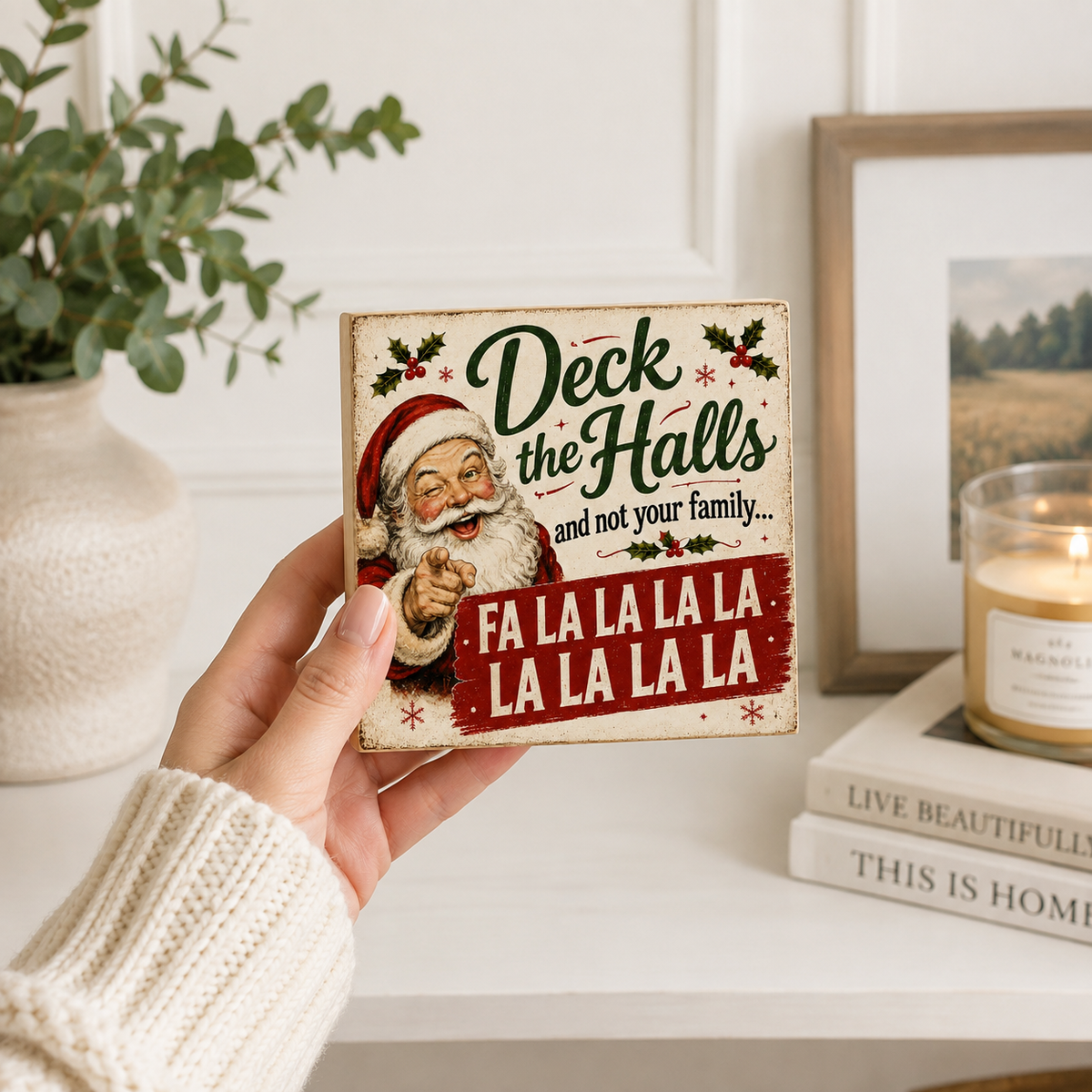 A hand in a cream knit sweater holds a square decorative Christmas sign that reads 'Deck the Halls and not your family.