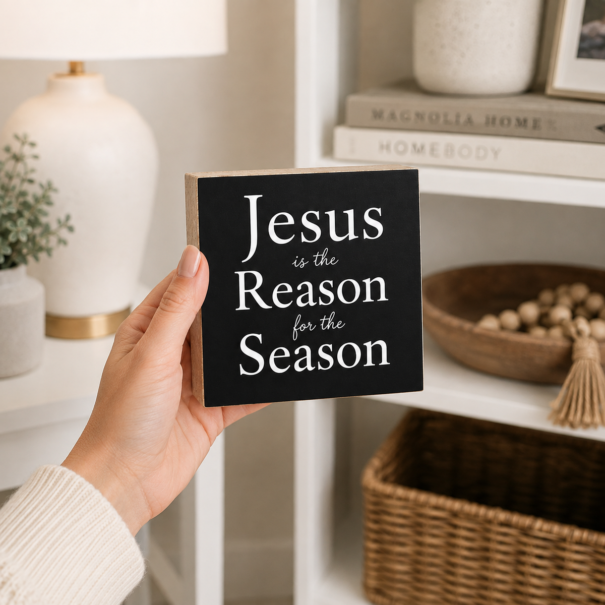 A hand holds a square black decorative sign with white text reading 'Jesus is the Reason for the Season' in a cozy, styled home interior setting.