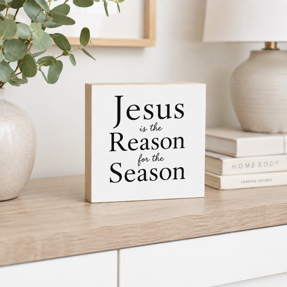 A square white decorative wood block sign with black text reading 'Jesus is the Reason for the Season' displayed on a light wood surface in a styled interior setting.