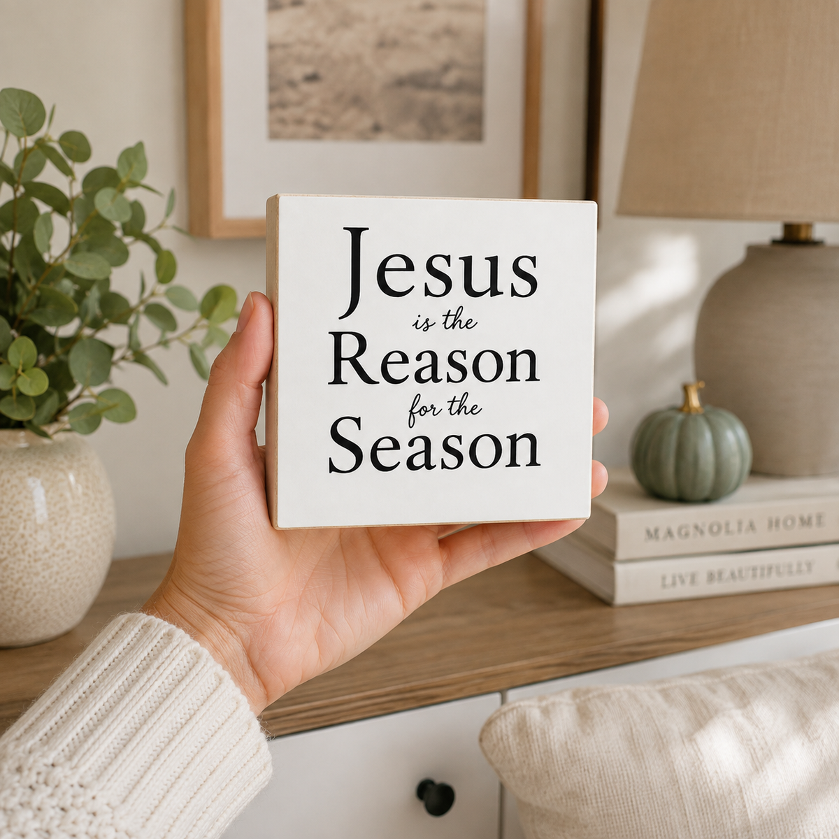 A hand holds a square white decorative sign with the text 'Jesus is the Reason for the Season' in black serif and script lettering. The background shows a cozy home interior with a lamp, books, a small pumpkin, and a potted plant.