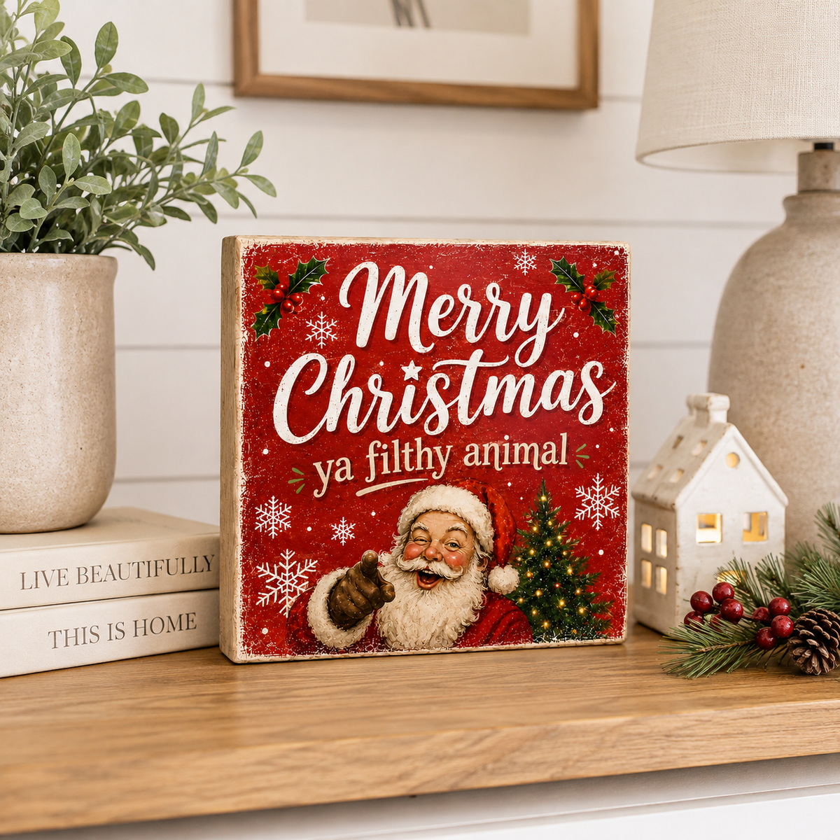A square Christmas sign with a red background, white text reading 'Merry Christmas ya filthy animal,' and an illustrated Santa pointing outward, styled on a wooden shelf with books, a plant, a lamp, and a small house decor piece.
