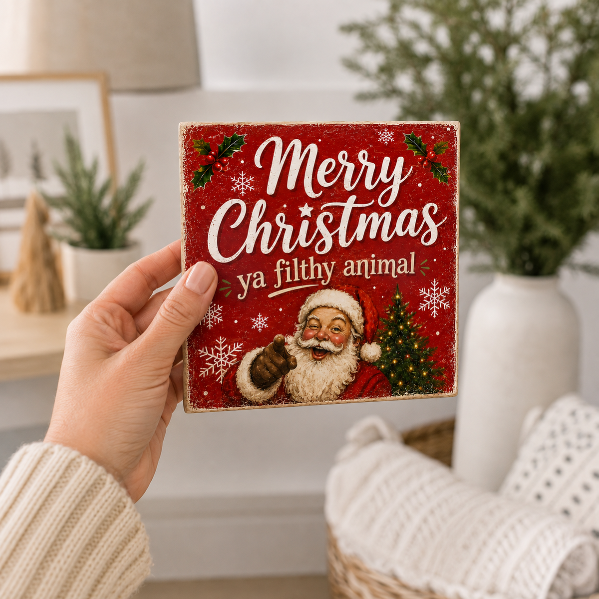 A hand holds a square red Christmas sign with a rustic wood-look finish. The sign features the text 'Merry Christmas ya filthy animal' in white, along with an illustration of Santa Claus, a small Christmas tree, and white snowflakes.
