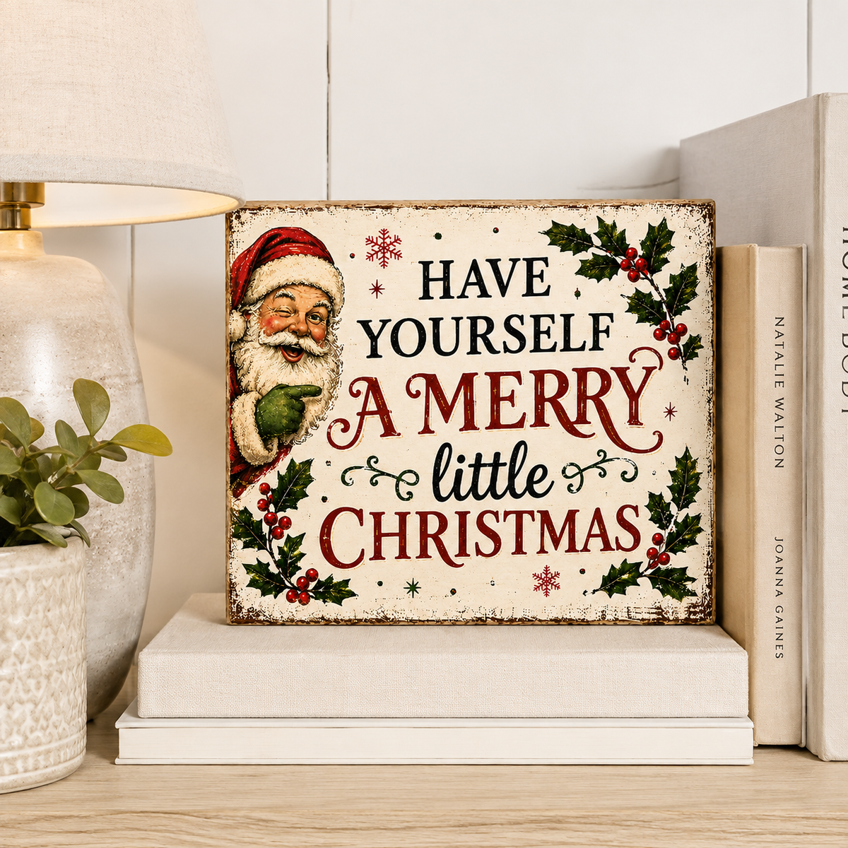 A rectangular Christmas sign with a vintage-style Santa illustration and the text 'HAVE YOURSELF A MERRY little CHRISTMAS' sits on a shelf beside a lamp, plant, and books.