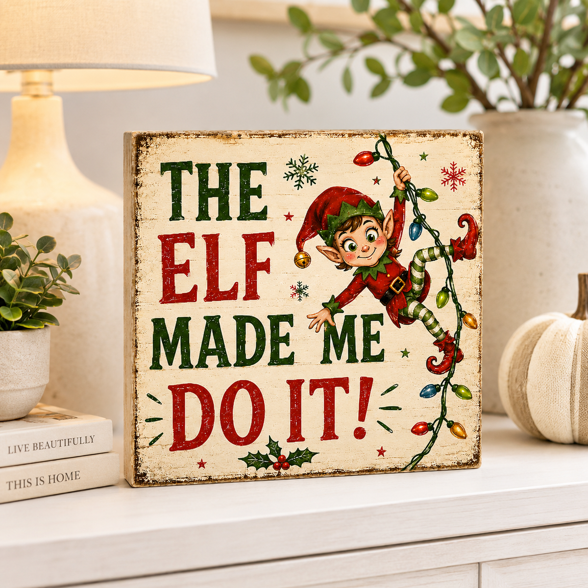 A rustic wooden Christmas sign with the phrase “THE ELF MADE ME DO IT!” in green and red lettering, featuring an illustrated elf climbing a string of multicolored lights. The sign is styled on a white shelf with a lamp, books, and a small pumpkin in the background.
