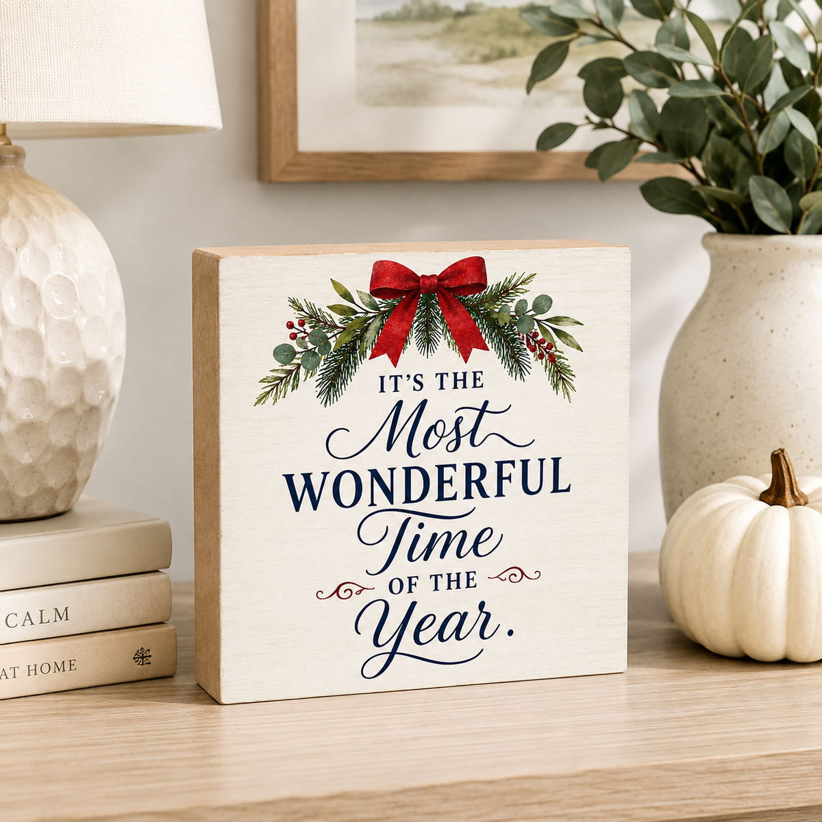 A square wooden sign with the phrase 'IT'S THE Most WONDERFUL Time OF THE Year.' in navy script and serif lettering, decorated with a red bow, green eucalyptus leaves, and red berries at the top.