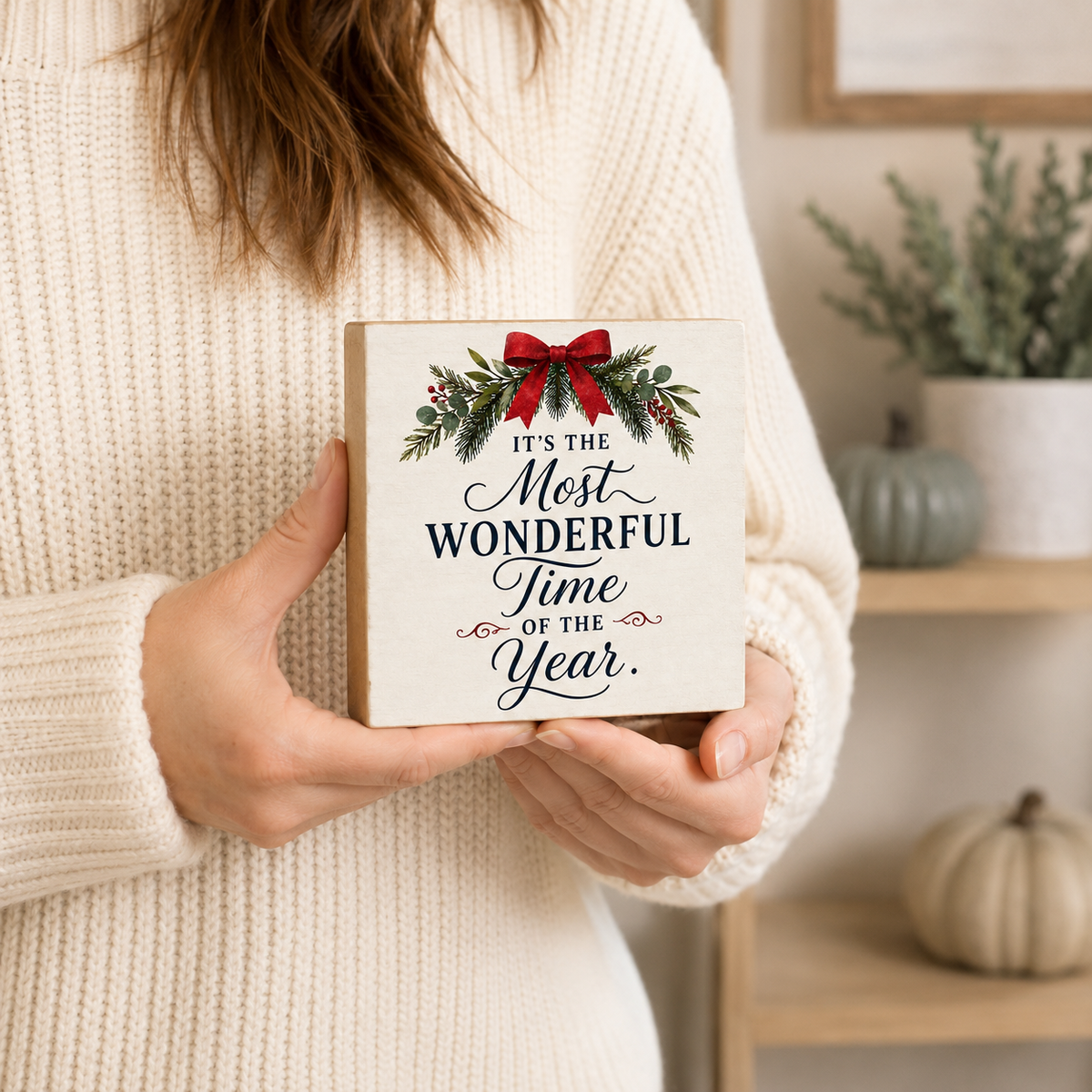 A person in a cream knit sweater holds a square wooden sign that reads 'It's the Most Wonderful Time of the Year.' The sign has a red bow and green pine garland illustration at the top.