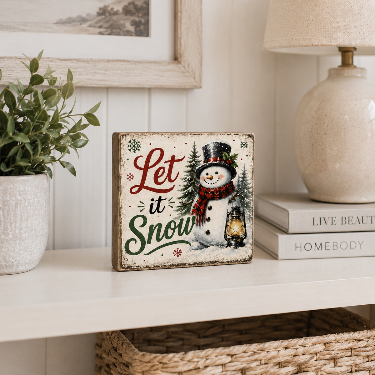 A square decorative sign with a distressed wood look featuring a snowman and the phrase 'Let it Snow' in red and green lettering, styled on a white shelf next to a potted plant, books, and a lamp.
