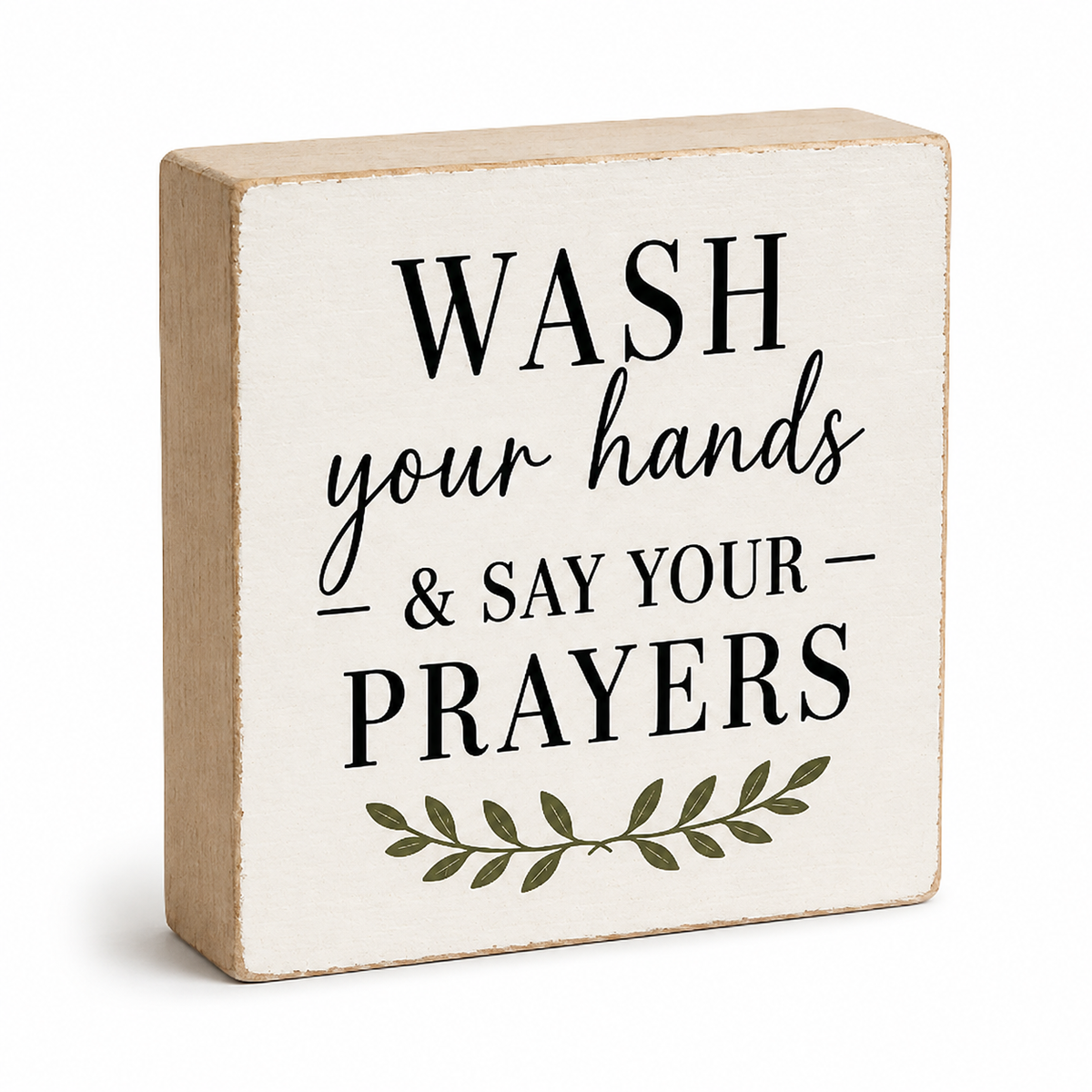 A square decorative wooden block sign with a distressed cream face and natural wood sides. The sign features the text 'WASH your hands & SAY YOUR PRAYERS' in black lettering, accented with a small green leafy branch graphic at the bottom, set against a plain white background.