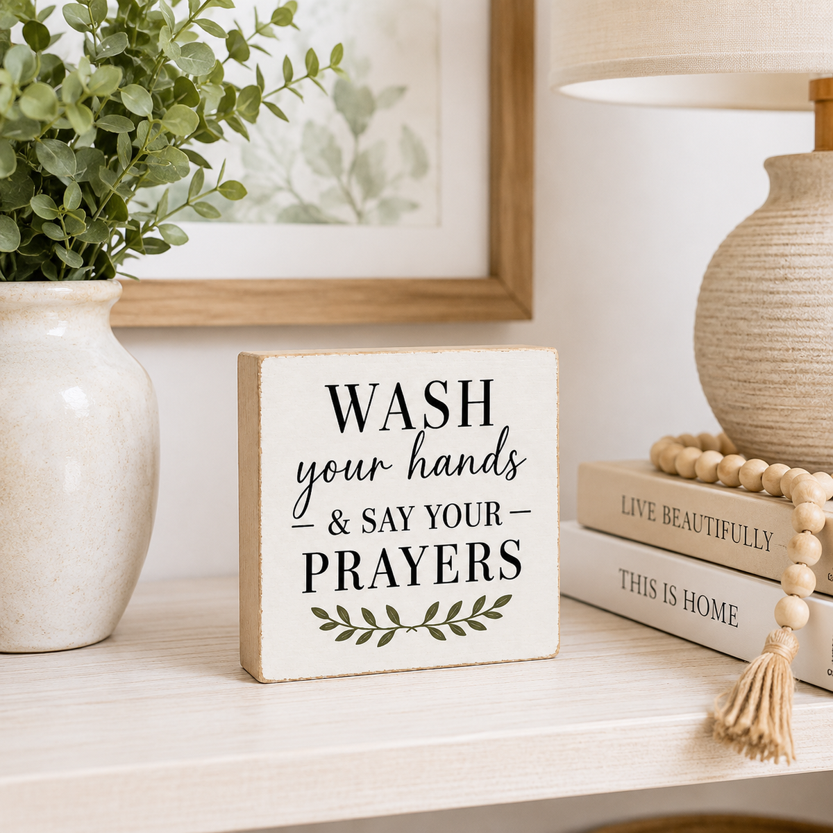 A square wooden sign with the text 'WASH your hands & SAY YOUR PRAYERS' and a small green leaf motif, styled on a tabletop beside a vase with greenery, books, a lamp, and a wooden bead tassel.
