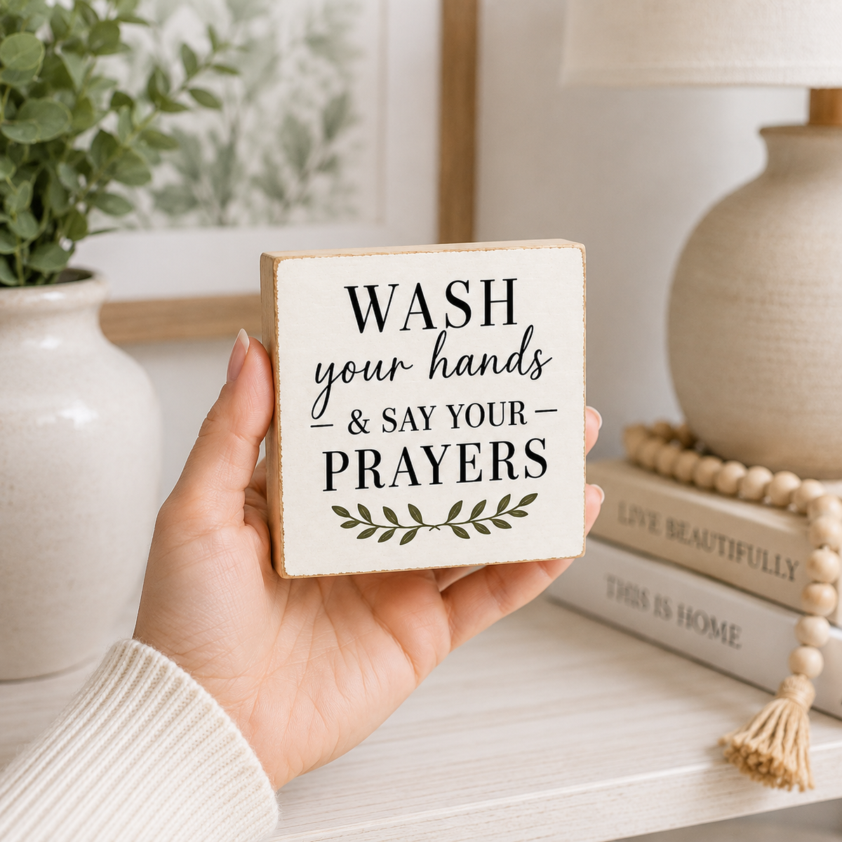 A hand holds a square wooden sign that reads 'WASH your hands & SAY YOUR PRAYERS' with a small leaf motif below. The background shows a softly styled home interior with a vase, framed art, a lamp, books, and a wooden bead necklace.