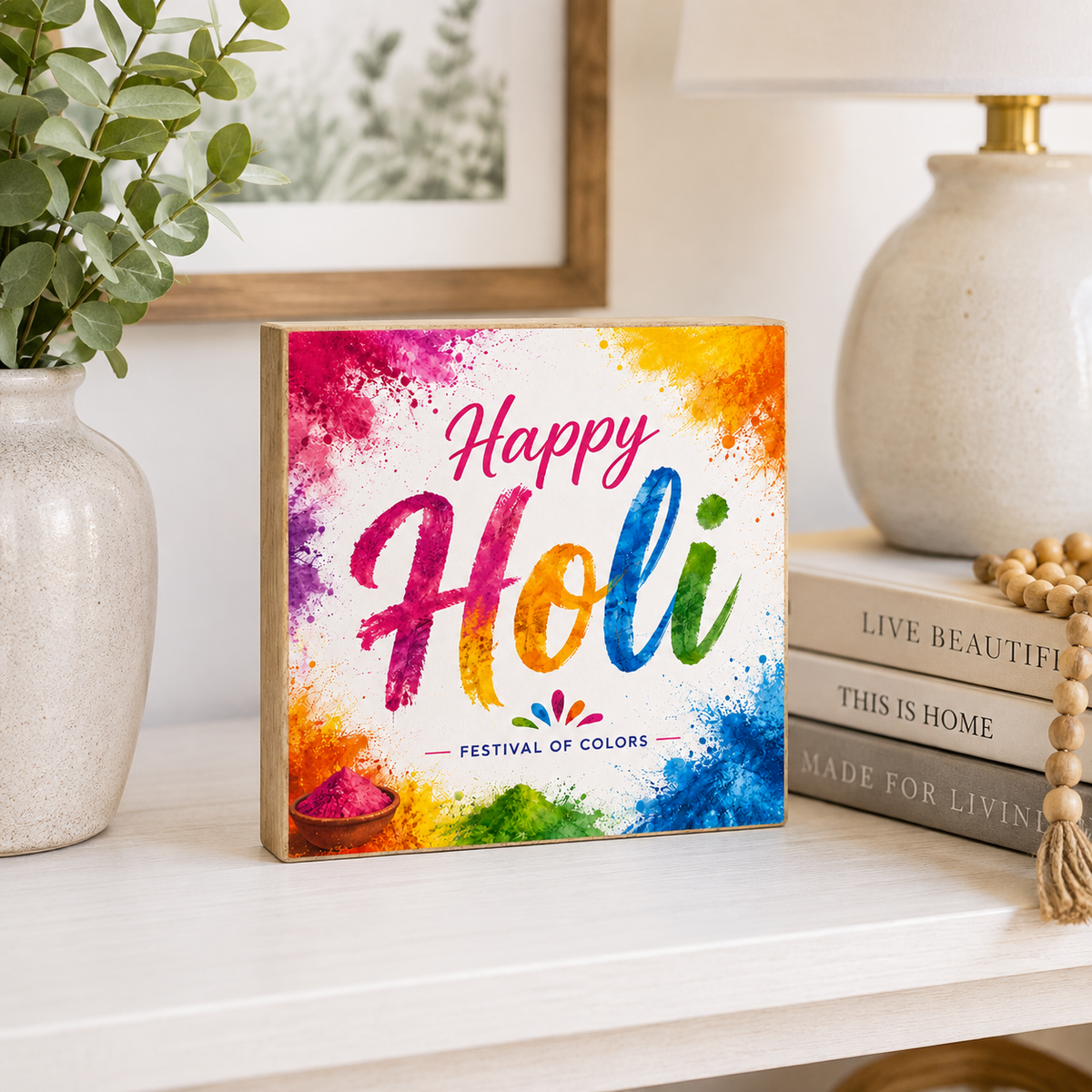 A square wooden sign with a colorful Holi watercolor design reading 'Happy Holi' and 'Festival of Colors,' styled on a white shelf beside a vase, framed art, a lamp, books, and a beaded necklace.
