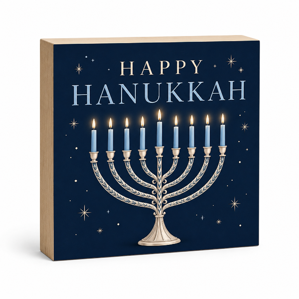 A square decorative block or plaque with a Hanukkah design, featuring the words “HAPPY HANUKKAH” in light blue and silver above a silver menorah with lit blue candles on a dark navy background with small gold stars. The block has a light wood side edge and is photographed on a clean white background.