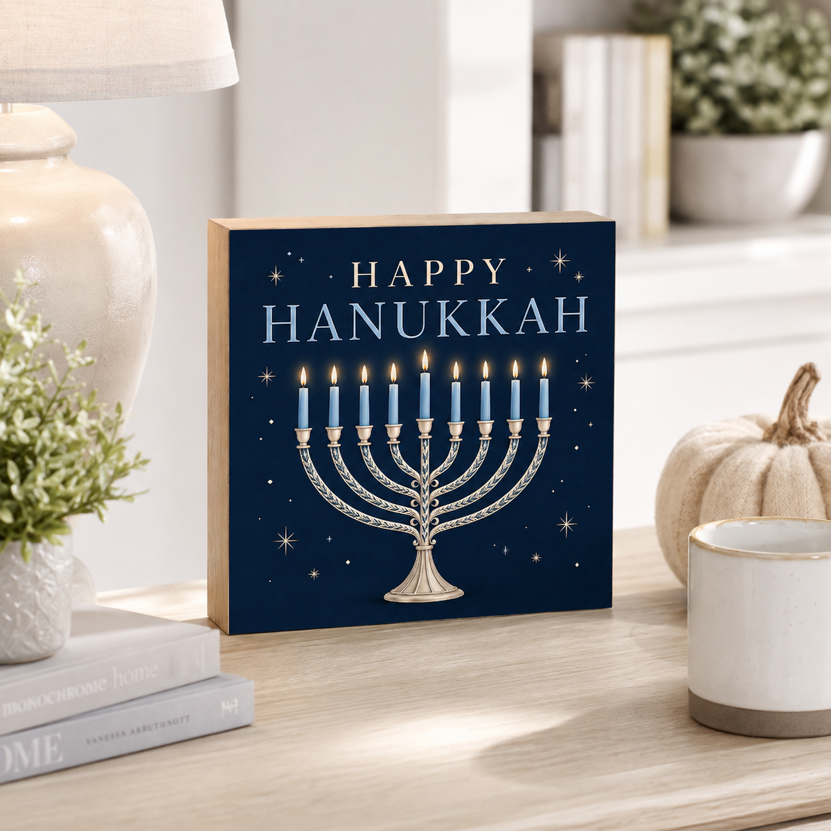 A square decorative sign with a Hanukkah design stands on a light wood tabletop in a styled home setting. The sign has a dark navy background with the words “HAPPY HANUKKAH” in white and a silver menorah with blue candles and small gold stars.