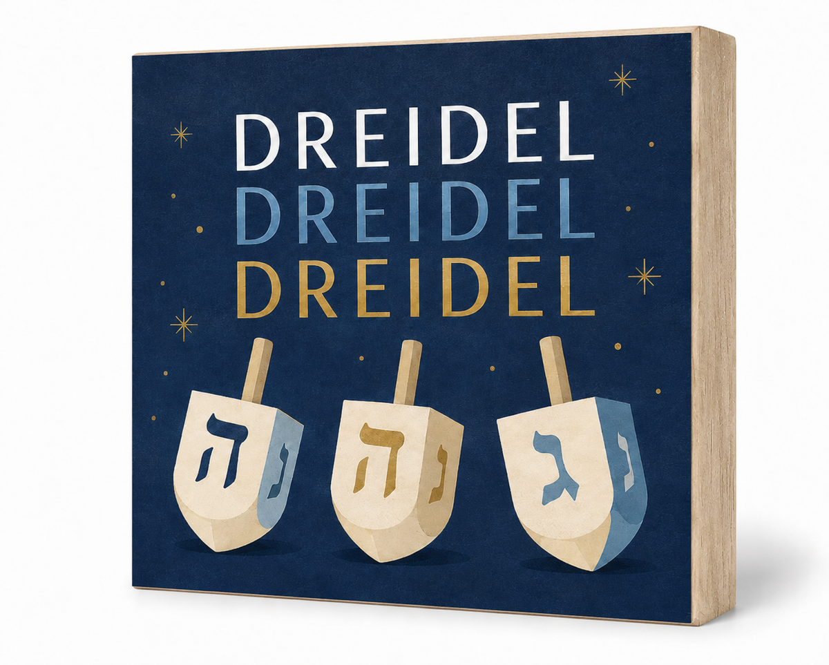 A square decorative sign or box front featuring the word 'DREIDEL' repeated three times in white, light blue, and gold text above three illustrated wooden dreidels on a dark navy background with small gold stars. The product is shown at a slight angle with a light wood side edge against a clean white background.