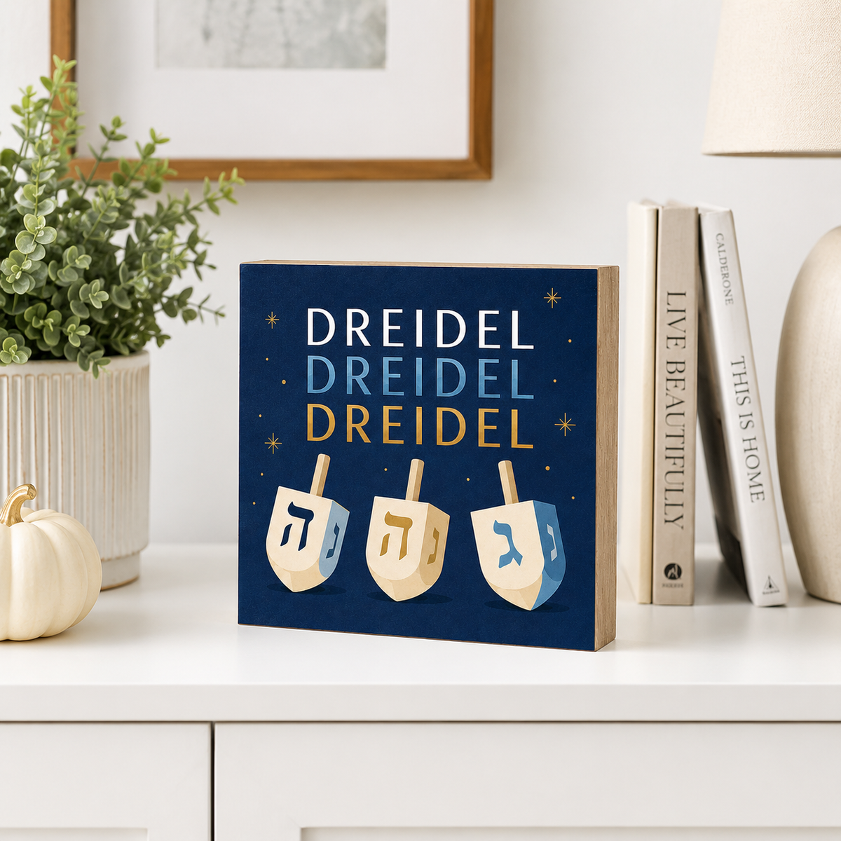 A square decorative sign featuring the word 'DREIDEL' repeated three times in white, light blue, and gold lettering above three illustrated dreidels, displayed on a white tabletop with books, a plant, and a small pumpkin nearby.