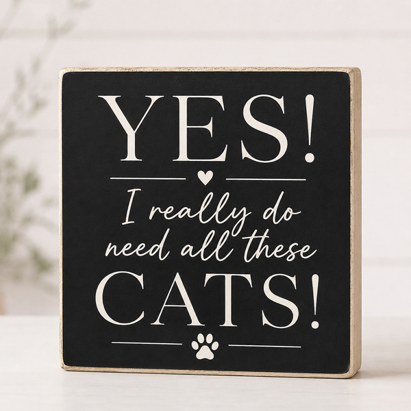 A square decorative wooden sign with a black face and natural wood edges, printed with the phrase “YES! I really do need all these CATS!” in white lettering, with a small paw print at the bottom, shown on a light surface.