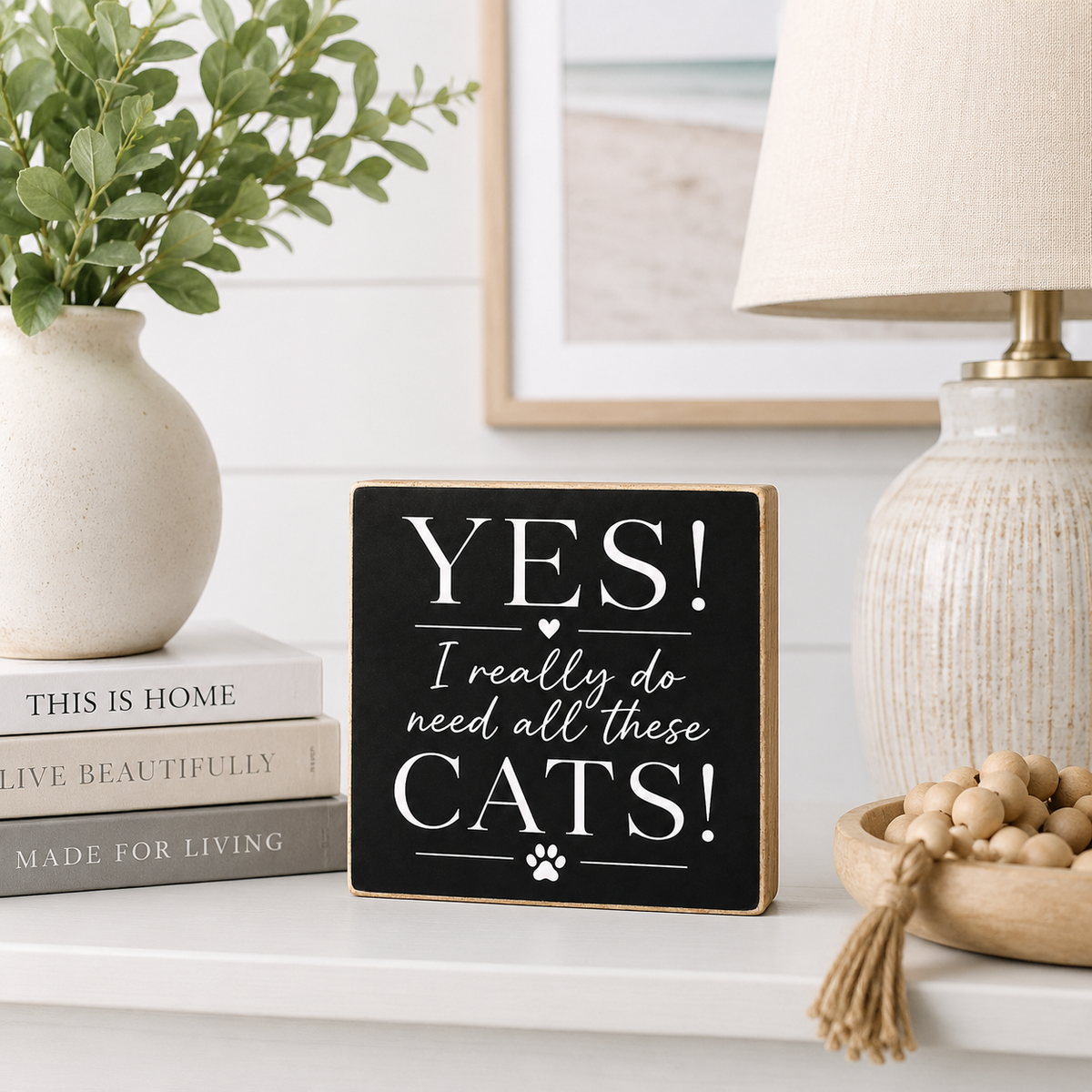 A square black decorative sign with a light wood border and white text reading “YES! I really do need all these CATS!” sits on a white shelf in a styled home setting with a vase of greenery, books, a lamp, and a bowl of beads.