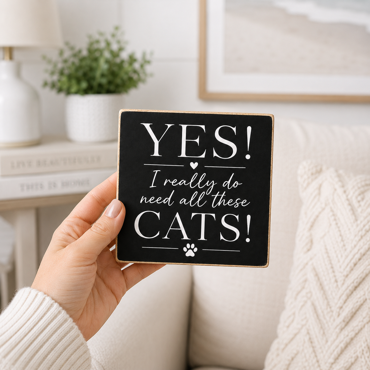 A hand holds a square black decorative sign with white text that reads “YES! I really do need all these CATS!” with a small paw print at the bottom. The background shows a cozy living room setting with a cream couch, a potted plant, and a lamp.