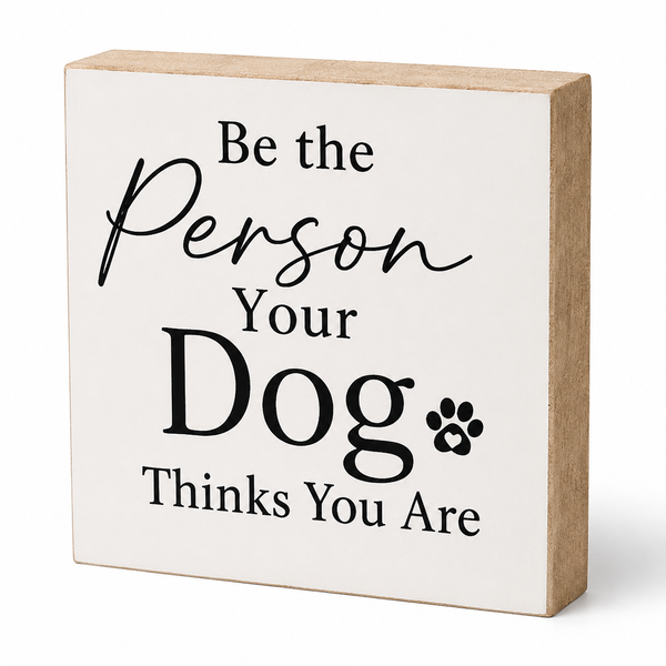 A square wooden sign with a white front panel and natural wood sides, featuring the printed phrase 'Be the Person Your Dog Thinks You Are' in black lettering with a small paw print.