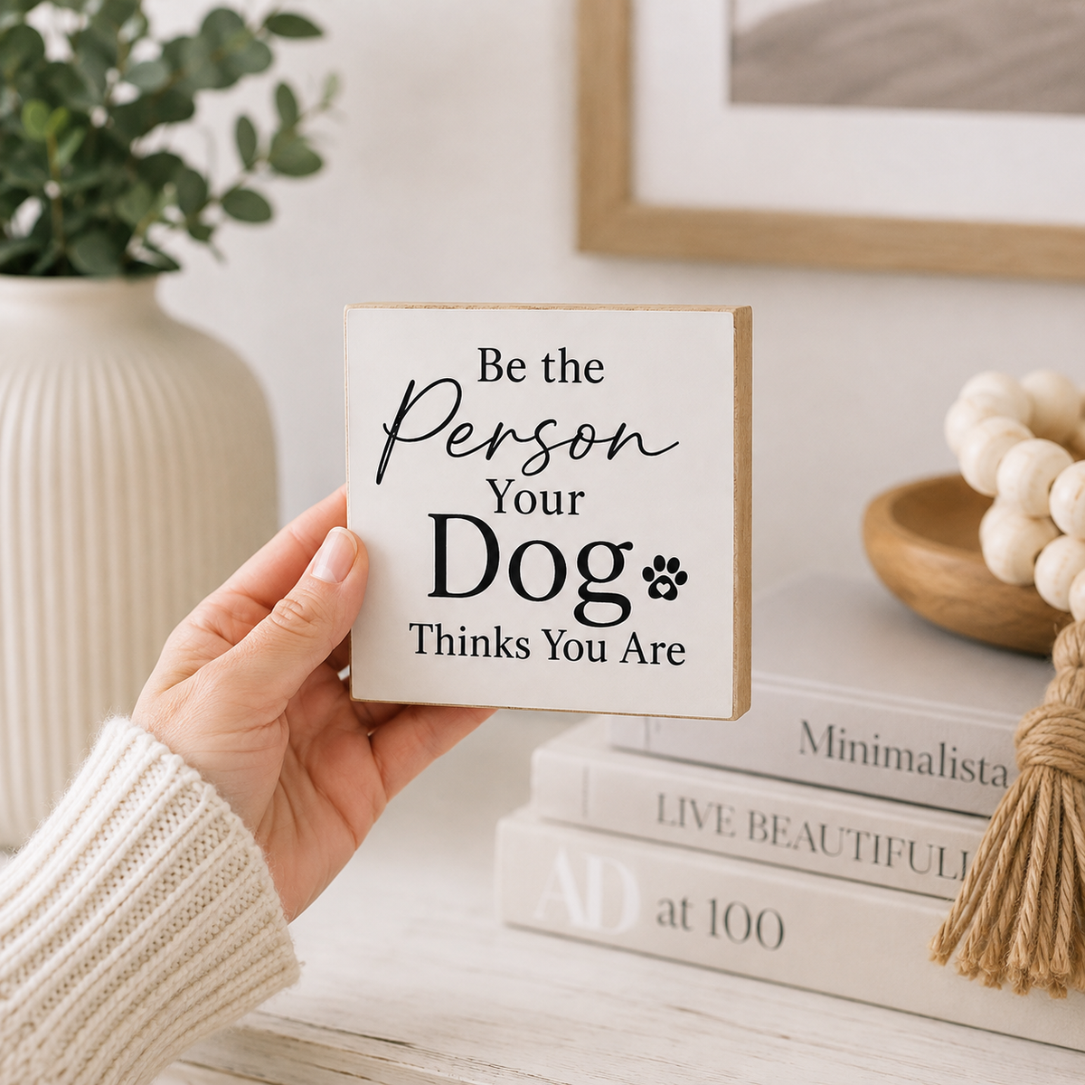 A hand wearing a cream knit sweater holds a square wooden sign that reads 'Be the Person Your Dog Thinks You Are' in black script and serif lettering. The background shows a softly styled home interior with a vase of greenery, framed wall art, stacked books, and a tassel decor piece.
