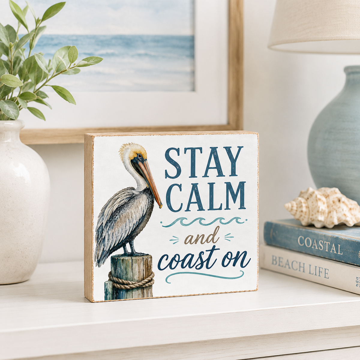 A square decorative sign with a painted pelican on a post and the phrase “STAY CALM and coast on,” styled on a shelf beside a vase, framed art, a lamp, and stacked books.
