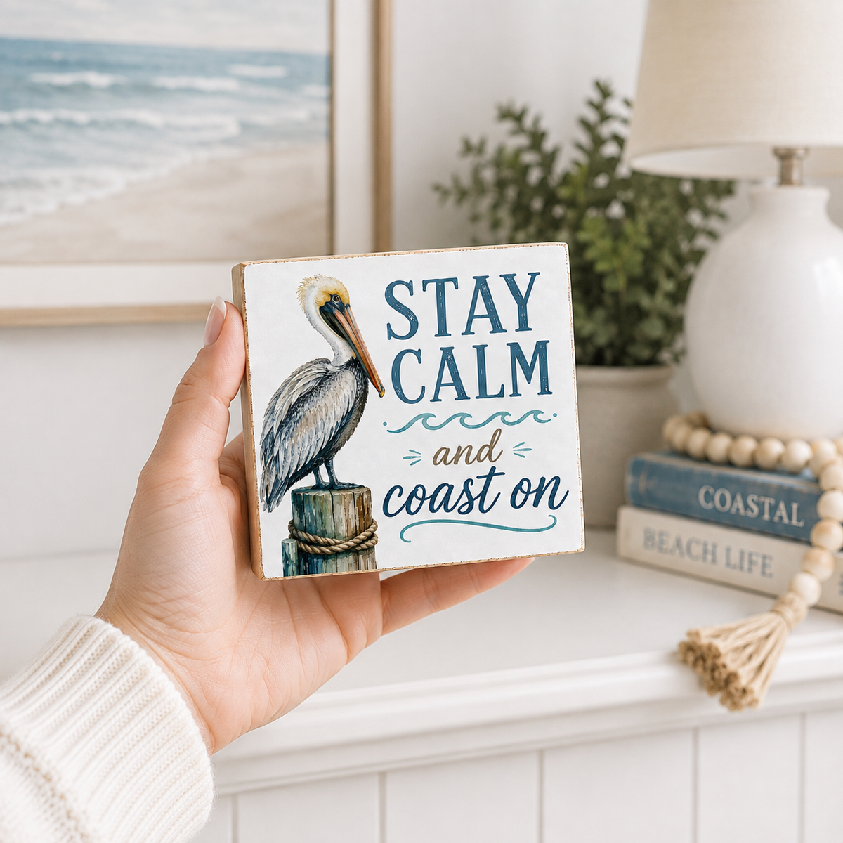 A hand holds a square wooden sign featuring a painted pelican and the phrase “STAY CALM and coast on.” The sign is shown in a bright, coastal-themed room setting with a lamp, books, and framed beach art in the background.