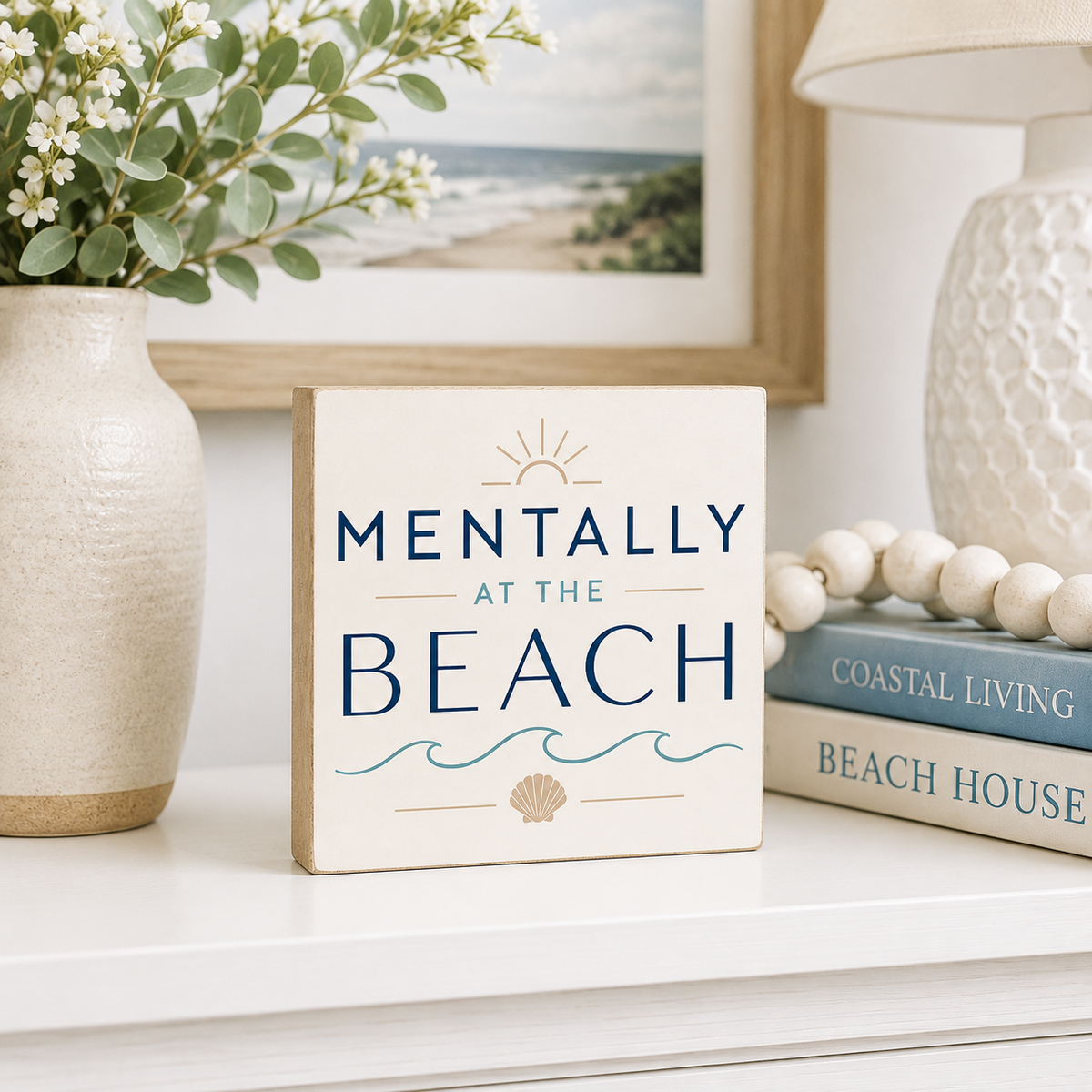 A square decorative sign reading “MENTALLY AT THE BEACH” in navy lettering with a small sun, waves, and seashell motif, styled on a white shelf beside a vase with greenery, a framed beach photo, a lamp, and stacked books.