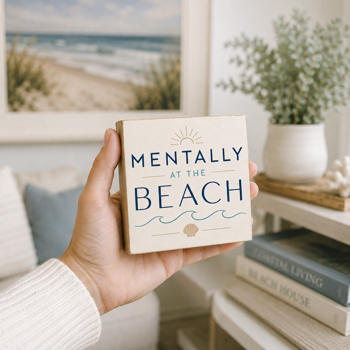 A hand holding a square cream-colored wooden sign that reads 'MENTALLY AT THE BEACH' in navy blue lettering, with a sun, waves, and seashell graphic. The background shows a cozy, softly blurred living room setting with a framed beach print, a potted plant, and books on a shelf.