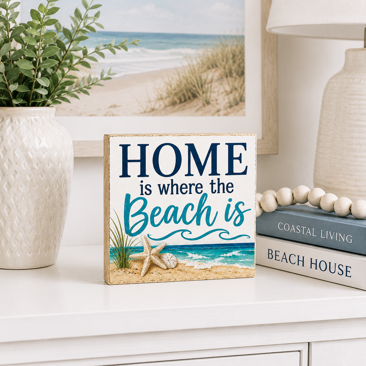 A square decorative sign with a beach scene and the text 'HOME is where the Beach is' displayed on a white dresser in a styled room setting.