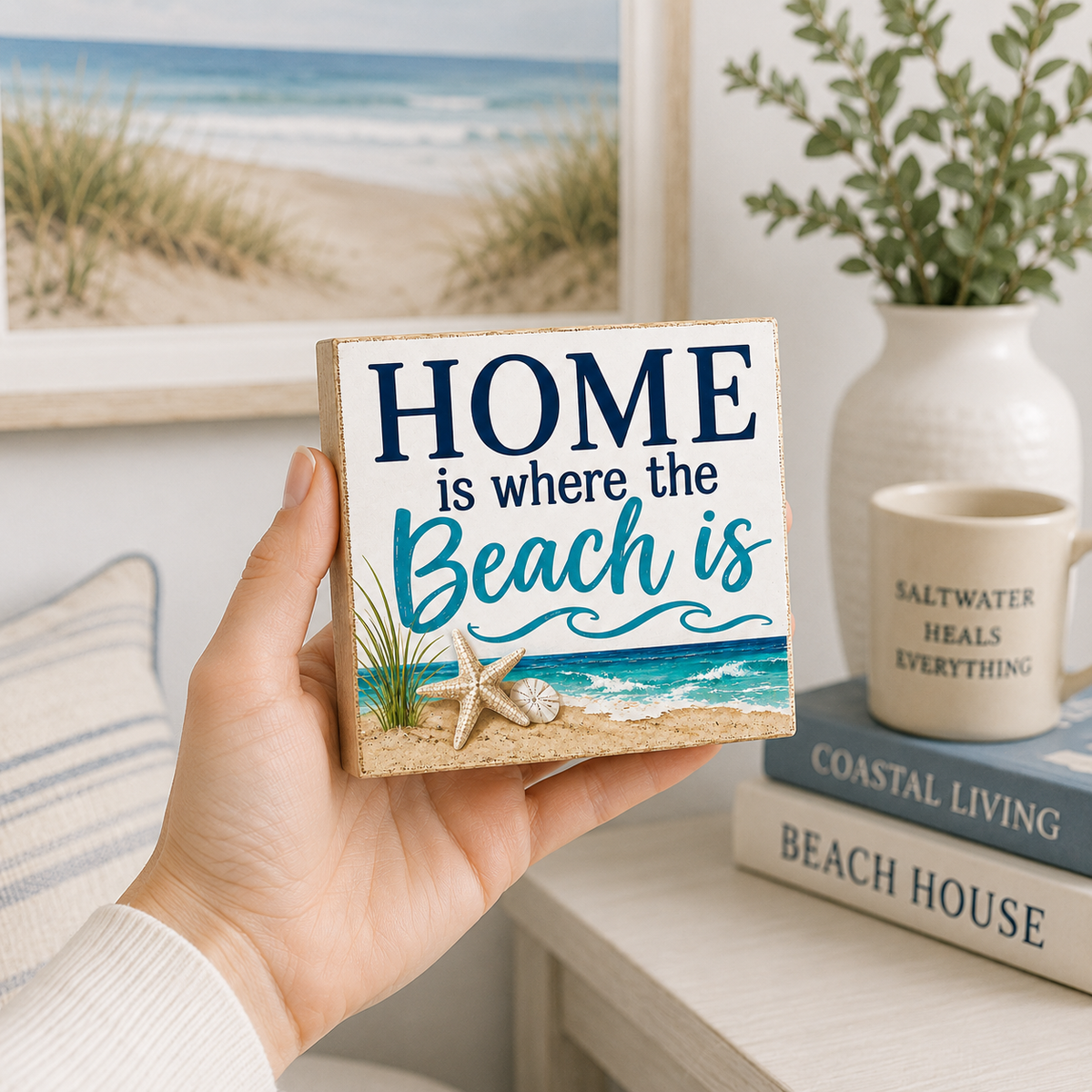 A hand holds a square wooden sign with the phrase 'HOME is where the Beach is' in navy and teal lettering above a beach scene with sand, grass, and a starfish. The background shows a styled interior with a framed beach print, a vase with greenery, a mug, and books.