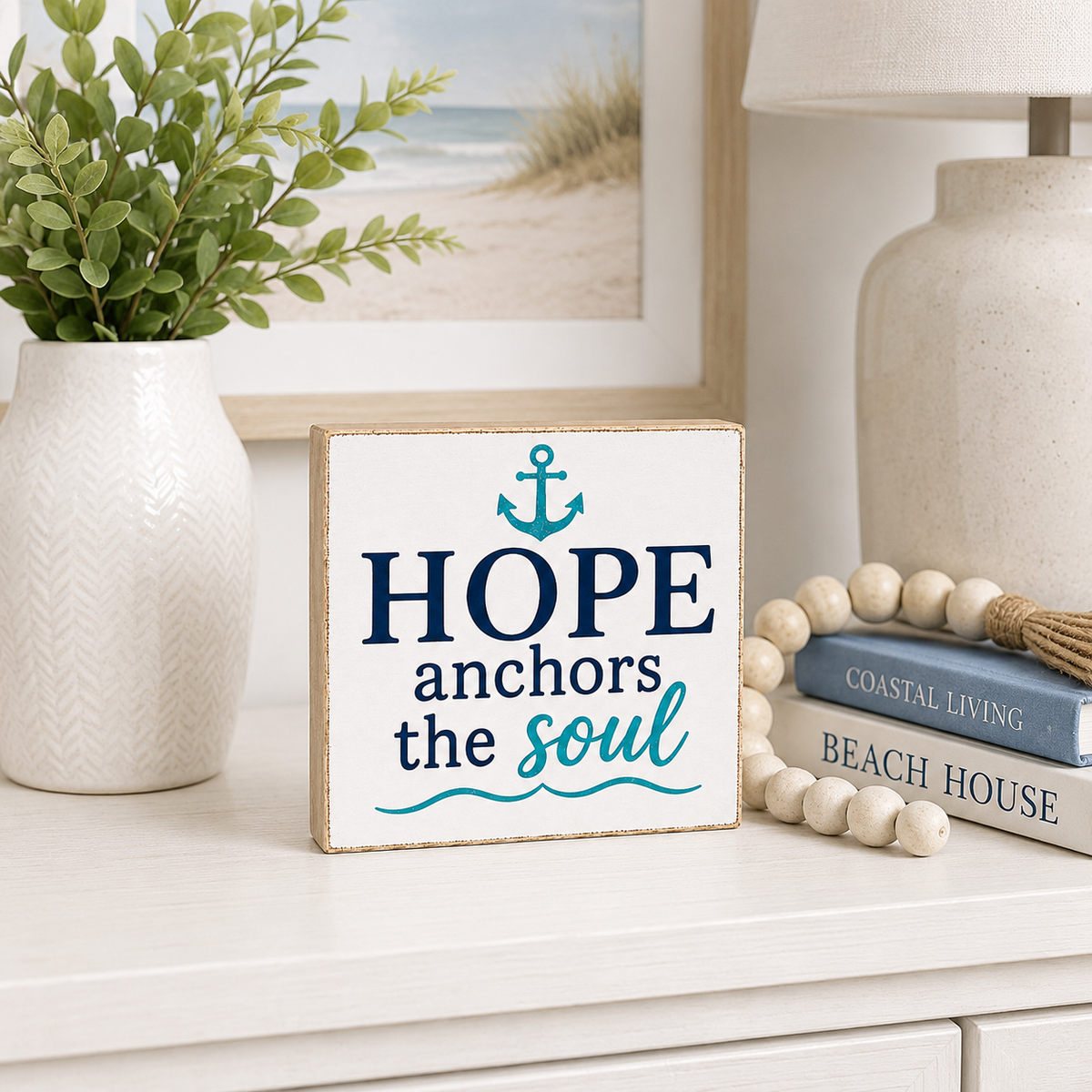 A square decorative sign with the phrase “HOPE anchors the soul” in navy and teal lettering on a white background, styled on a white tabletop with a vase, framed beach print, lamp, books, and wooden beads.